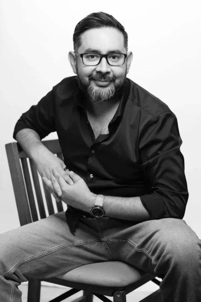 A man, Michael Samparia with glasses and a beard, wearing a dark shirt and jeans, sitting on a chair and smiling.